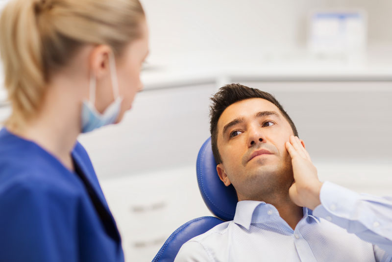a dental patient sitting in a procedure chair, while holding his jaw and talking to an oral surgeon about getting a comfortable wisdom tooth removal.